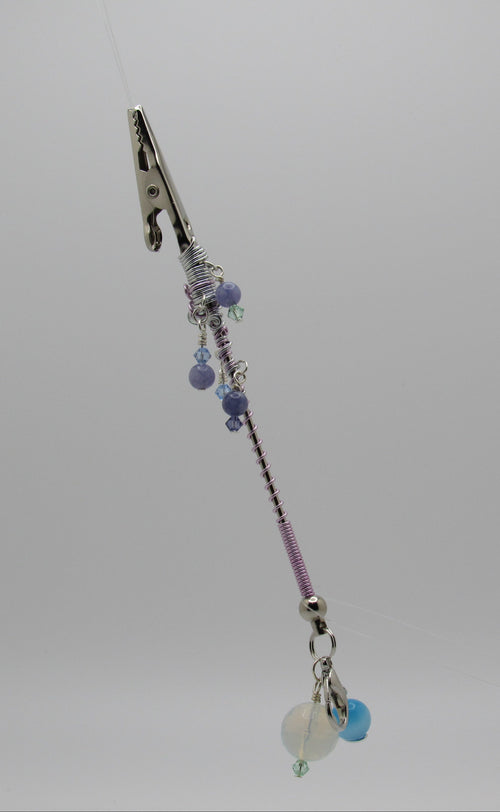 A beaded accessory with a silver metal clip at the top, purple and silver beads, and small dangling charms, shown against a plain light gray background.