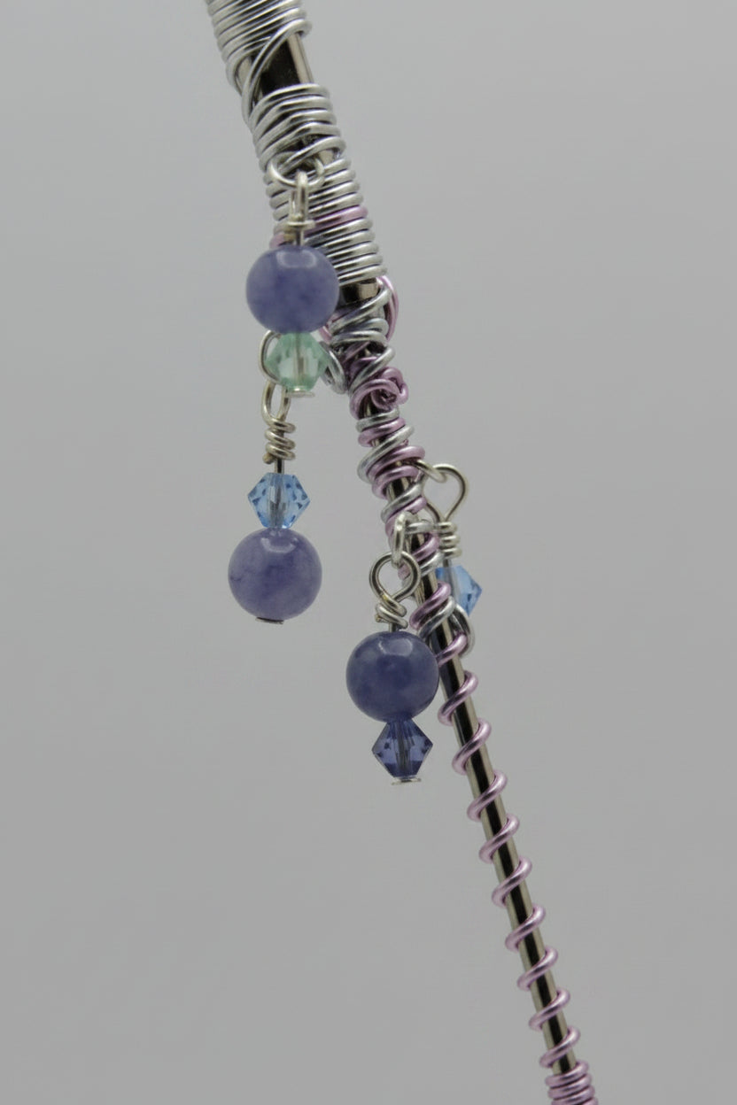 A close-up of a beaded roach clip with purple and blue beads, silver and lavender wire wrapping, and a silver metal shaft, set against a solid gray background.