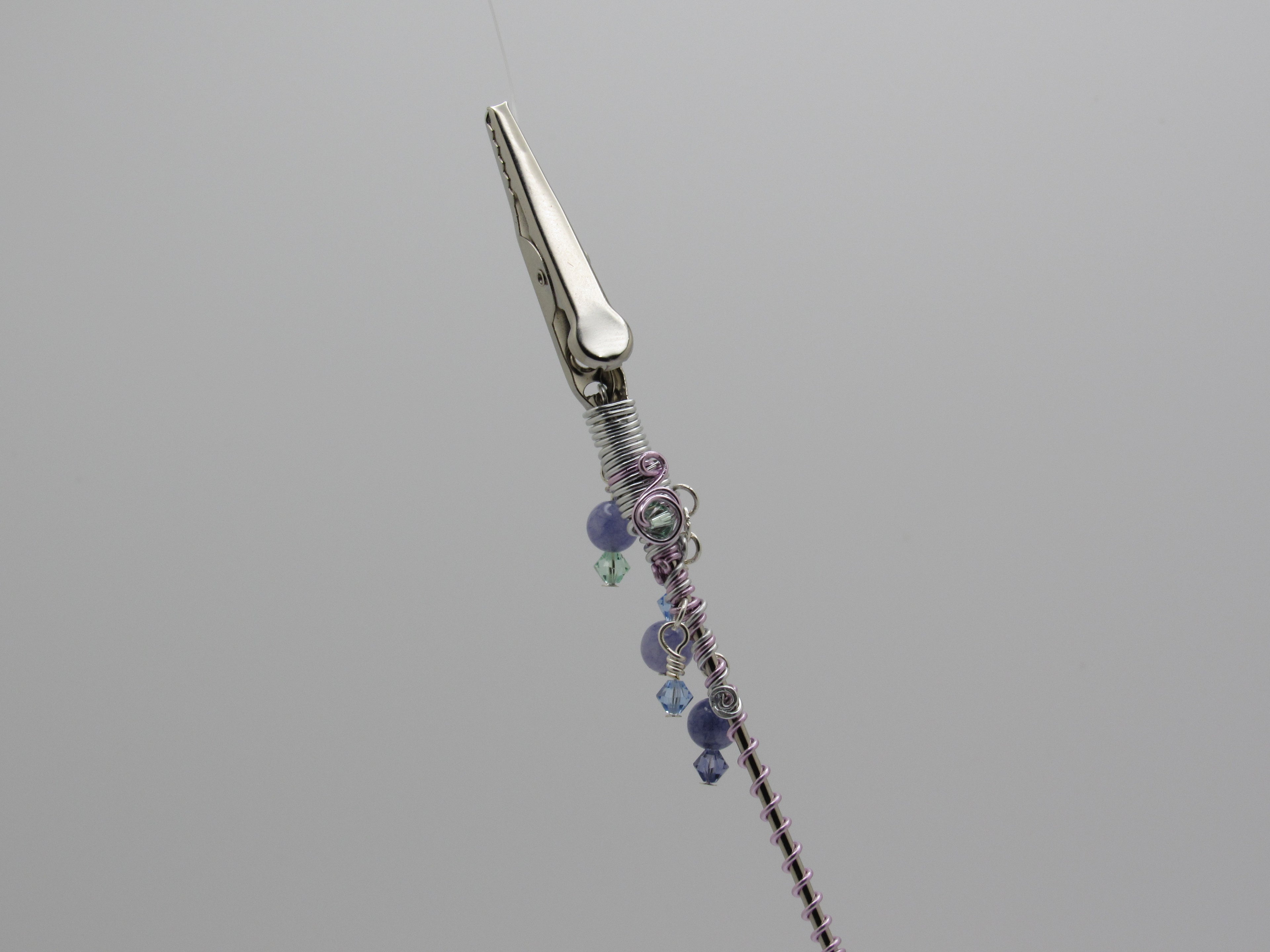 close up of top of roach clip, wrapped wire, and small purple and blue beads, shown against a solid gray background.