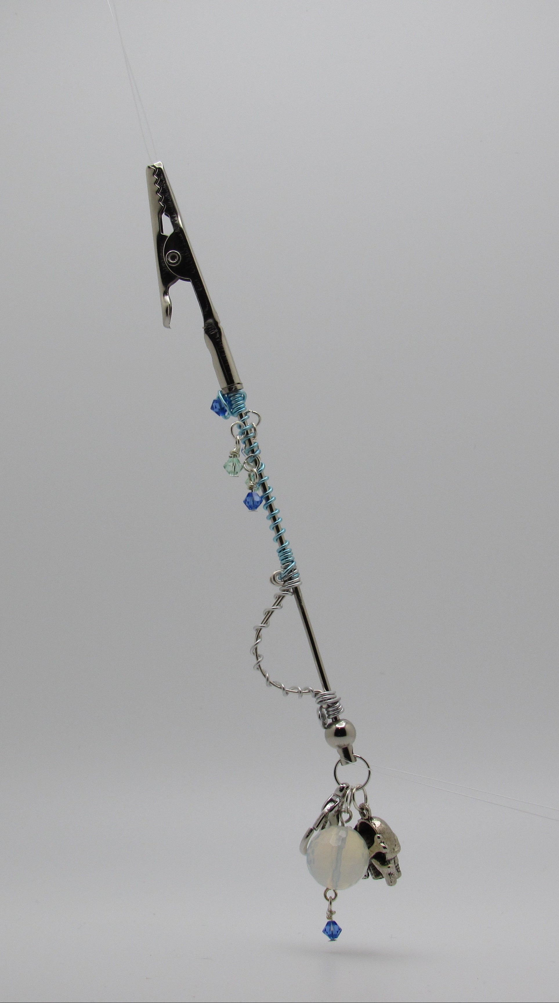 A silver-toned card roach clip with a metal clip at the top, blue beads, a chain, and a dangling silver bell with a small blue bead, photographed against a light gray studio background.