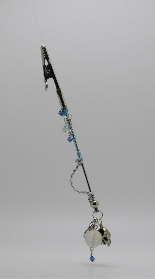 A silver-toned card roach clip with a metal clip at the top, blue beads, a chain, and a dangling silver bell with a small blue bead, photographed against a light gray studio background.