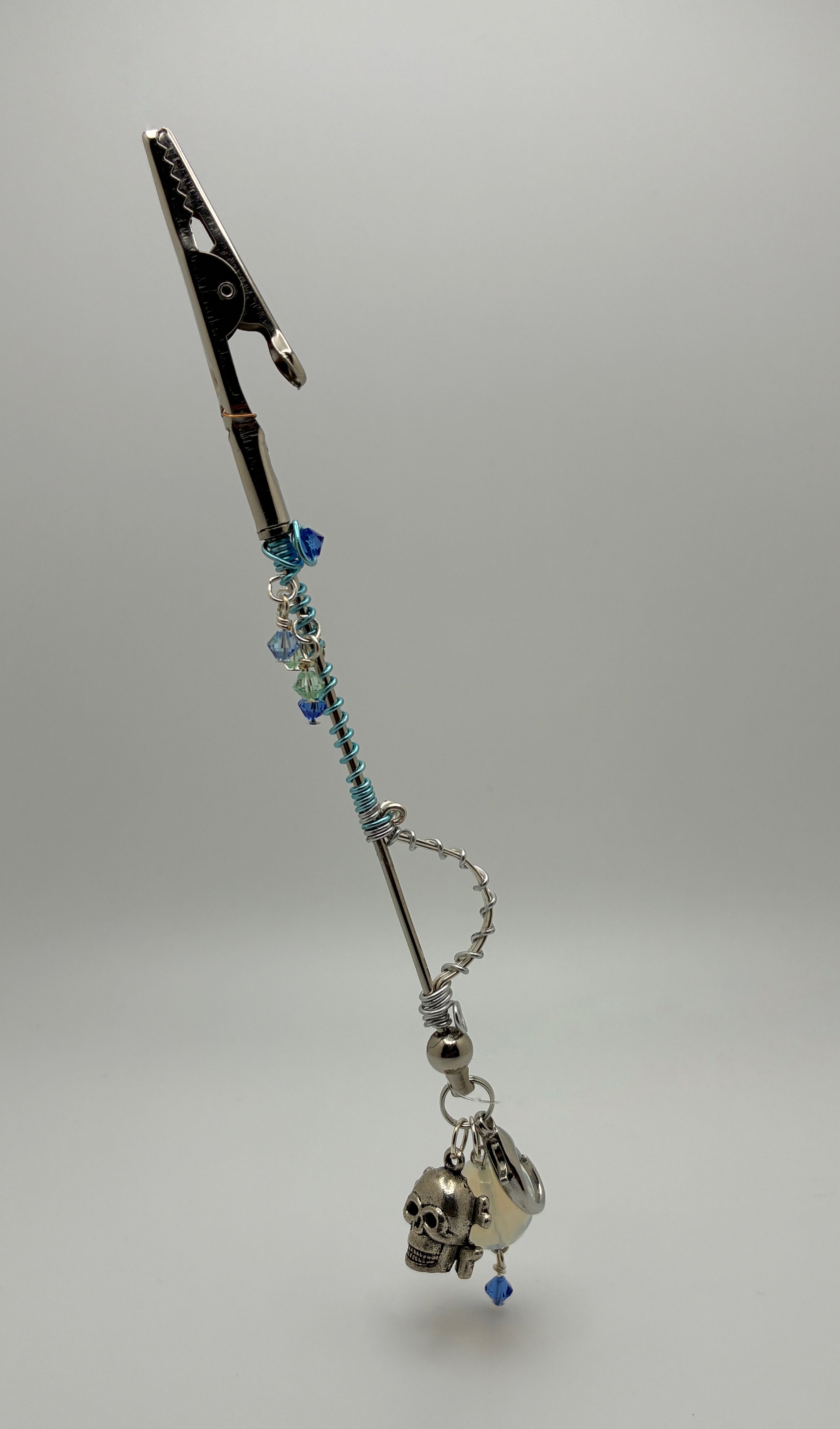 A gothic-style silver-toned car roach clip wand with blue beads, a skull charm, shown on a neutral gray background.