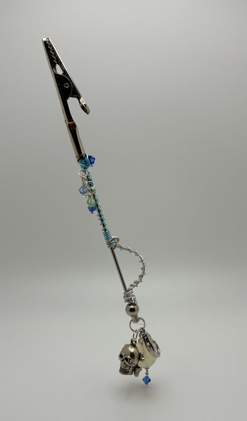 A gothic-style silver-toned car roach clip wand with blue beads, a skull charm, shown on a neutral gray background.